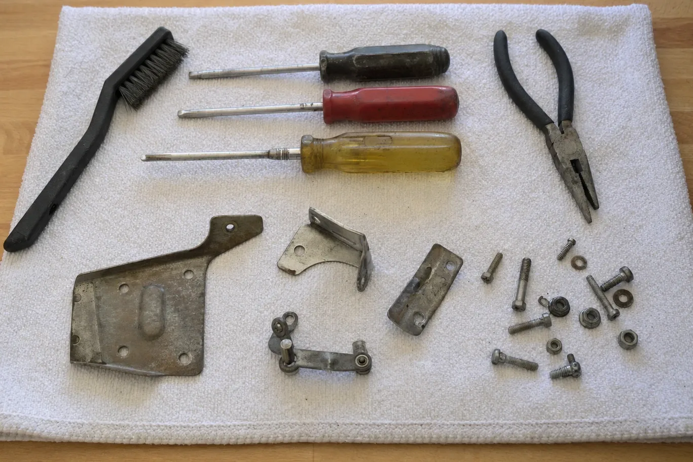 Three screwdrivers, pliers and a wire brush on a towel with bracket plates, a hinge link, bolts and nuts